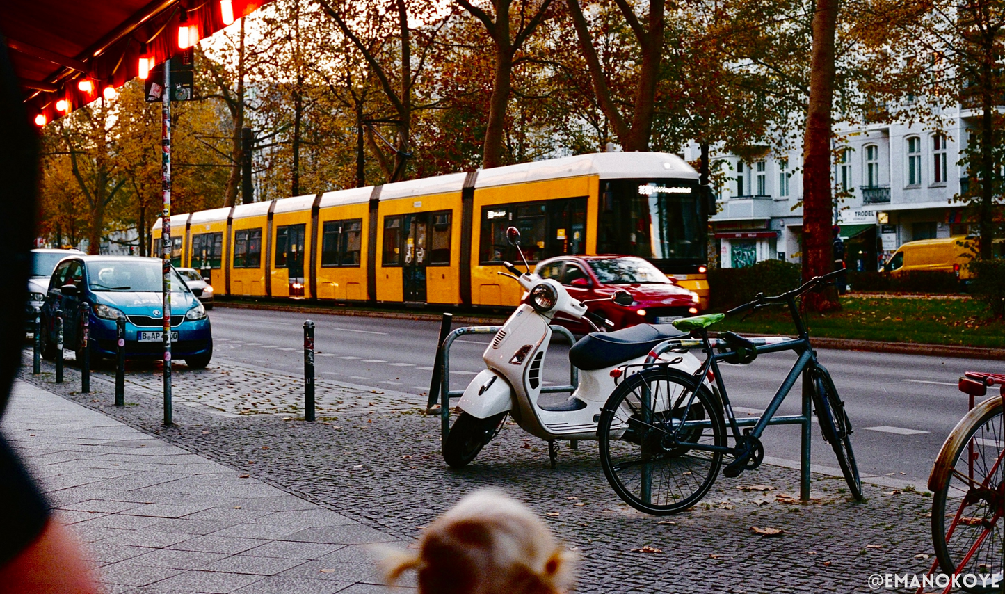 a vespa chained up to a bike rack with a yellow city tram in the background